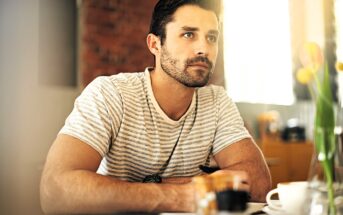 A man with short dark hair and a beard is sitting indoors, wearing a striped shirt. He gazes thoughtfully to the side, resting his arms on a table with a cup and a pot of colored pencils nearby. Tulips in a vase are partially visible.