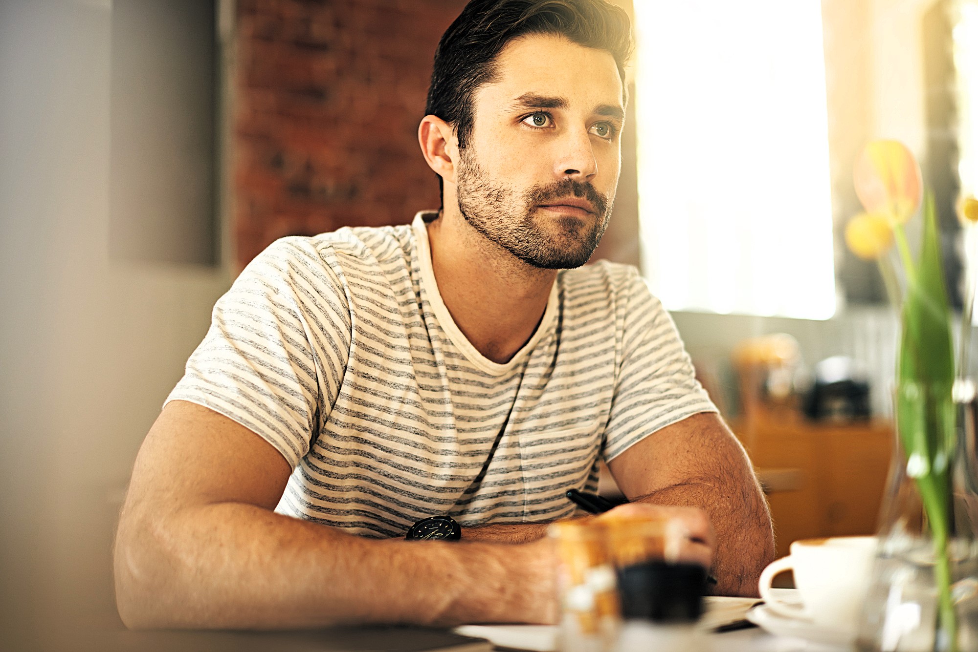 A man with short dark hair and a beard is sitting indoors, wearing a striped shirt. He gazes thoughtfully to the side, resting his arms on a table with a cup and a pot of colored pencils nearby. Tulips in a vase are partially visible.