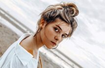 A woman with hair in a messy bun, wearing large hoop earrings and a white shirt, stands on a beach. Her intense gaze is directed at the camera. The ocean and an overcast sky serve as the background.