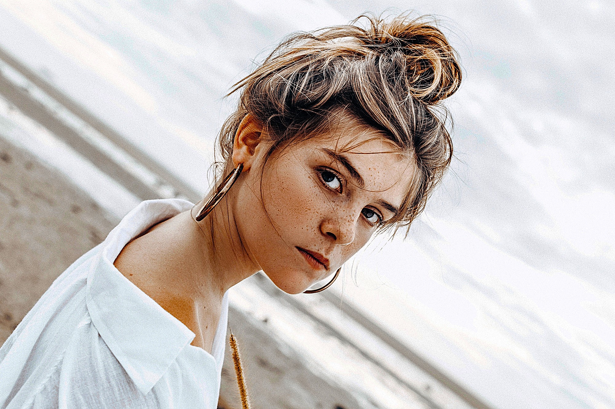 A woman with hair in a messy bun, wearing large hoop earrings and a white shirt, stands on a beach. Her intense gaze is directed at the camera. The ocean and an overcast sky serve as the background.