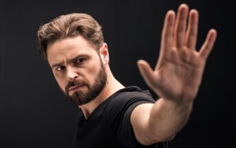 A man with a beard and short hair, wearing a black shirt, holds his hand in front of him with his palm facing forward. He appears focused and is looking in the direction of his hand. The background is plain and dark.