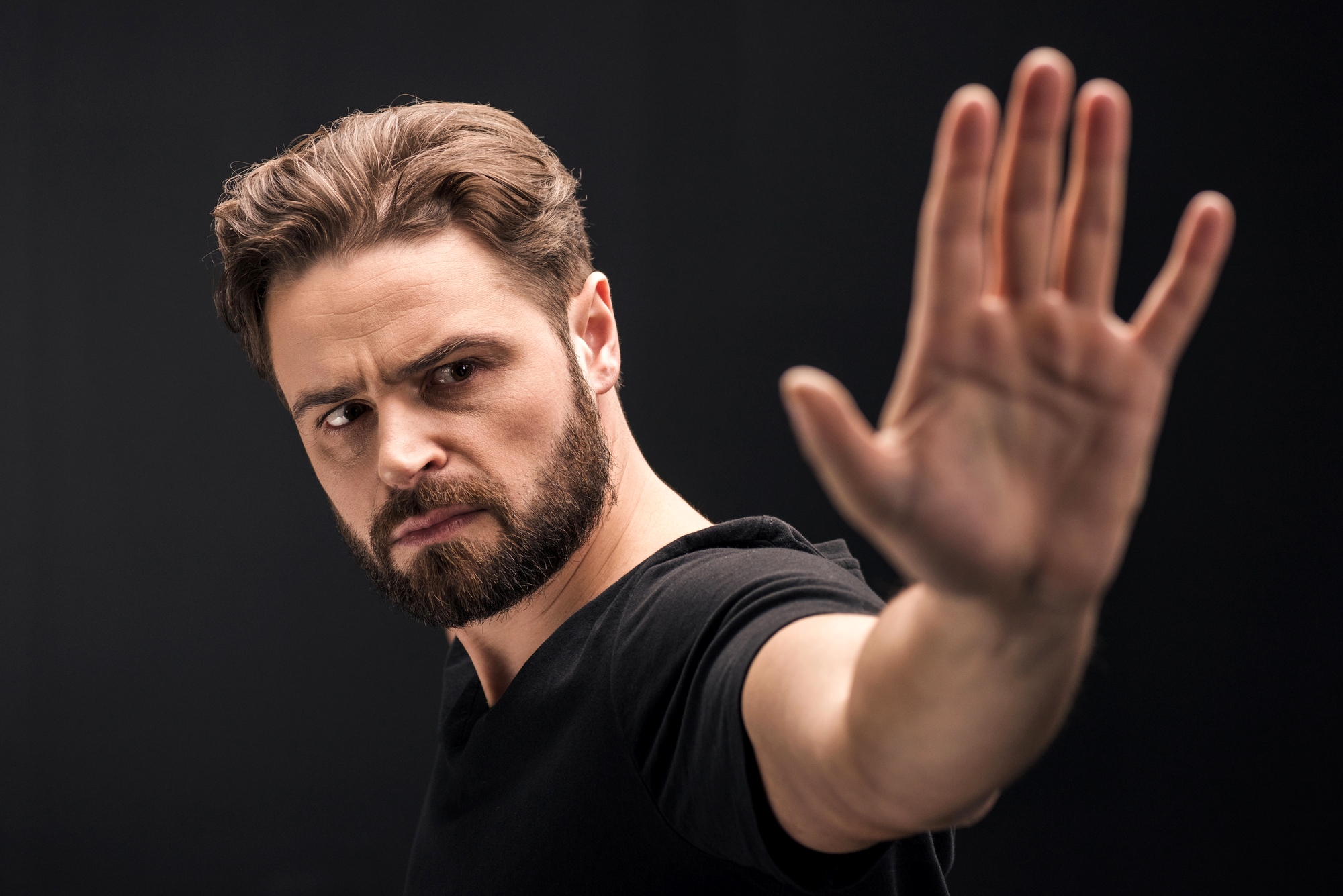 A man with a beard and short hair, wearing a black shirt, holds his hand in front of him with his palm facing forward. He appears focused and is looking in the direction of his hand. The background is plain and dark.