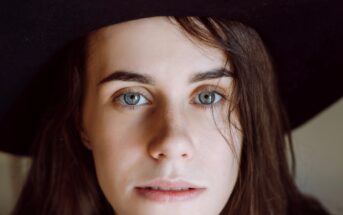 A close-up of a person with long brown hair and blue eyes, wearing a wide-brimmed black hat. They have a neutral expression, and one side of their face is partially covered by hair. The background is softly blurred.