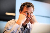 A man in a blue shirt sits at a desk, resting his face in his hands with a frustrated expression. He appears to be looking at a computer screen. The background is out of focus, suggesting an office environment.