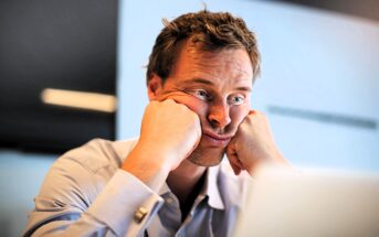 A man in a blue shirt sits at a desk, resting his face in his hands with a frustrated expression. He appears to be looking at a computer screen. The background is out of focus, suggesting an office environment.