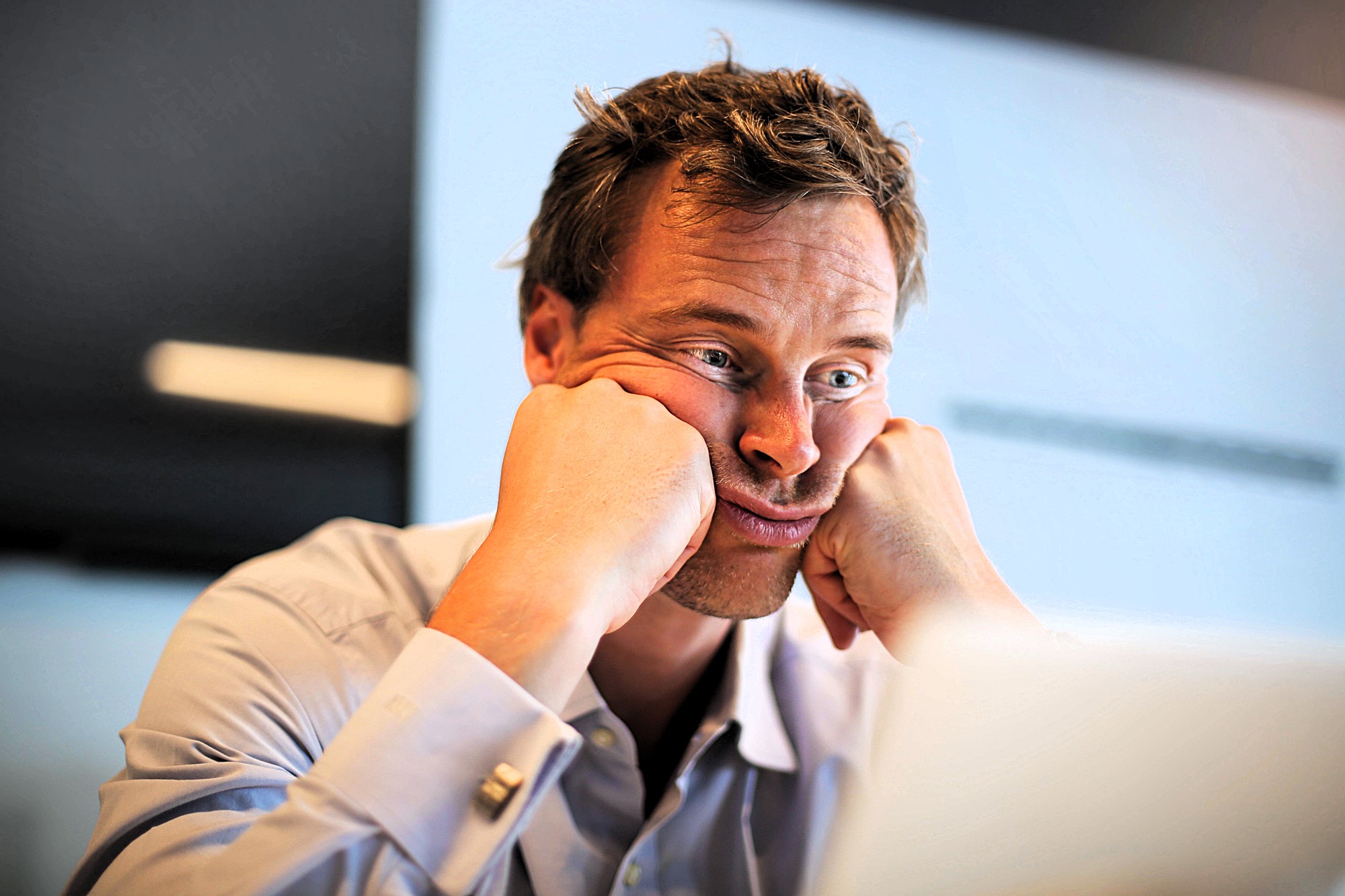 A man in a blue shirt sits at a desk, resting his face in his hands with a frustrated expression. He appears to be looking at a computer screen. The background is out of focus, suggesting an office environment.