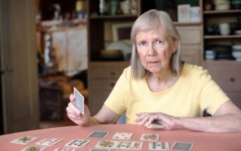 An elderly woman with gray hair and a yellow shirt is playing cards at a table. She is holding a card in her right hand, and several cards are laid out in front of her. She is sitting in a cozy room with shelves and cabinets in the background.