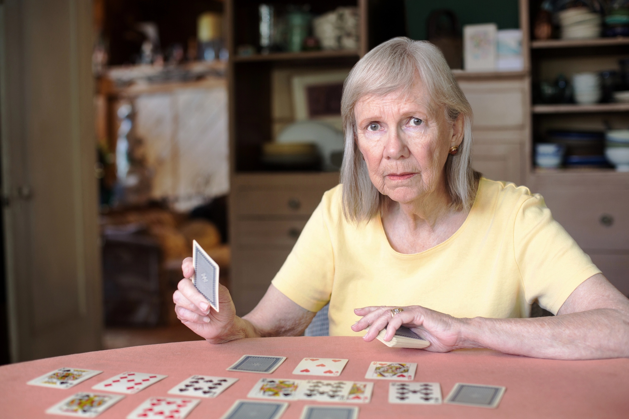 An elderly woman with gray hair and a yellow shirt is playing cards at a table. She is holding a card in her right hand, and several cards are laid out in front of her. She is sitting in a cozy room with shelves and cabinets in the background.