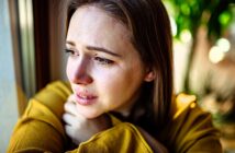 A woman with tearful eyes gazes out of a window, wearing a yellow sweater. She appears deep in thought, with sunlight illuminating her face and a blurred green background outside.