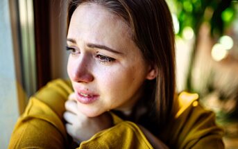 A woman with tearful eyes gazes out of a window, wearing a yellow sweater. She appears deep in thought, with sunlight illuminating her face and a blurred green background outside.