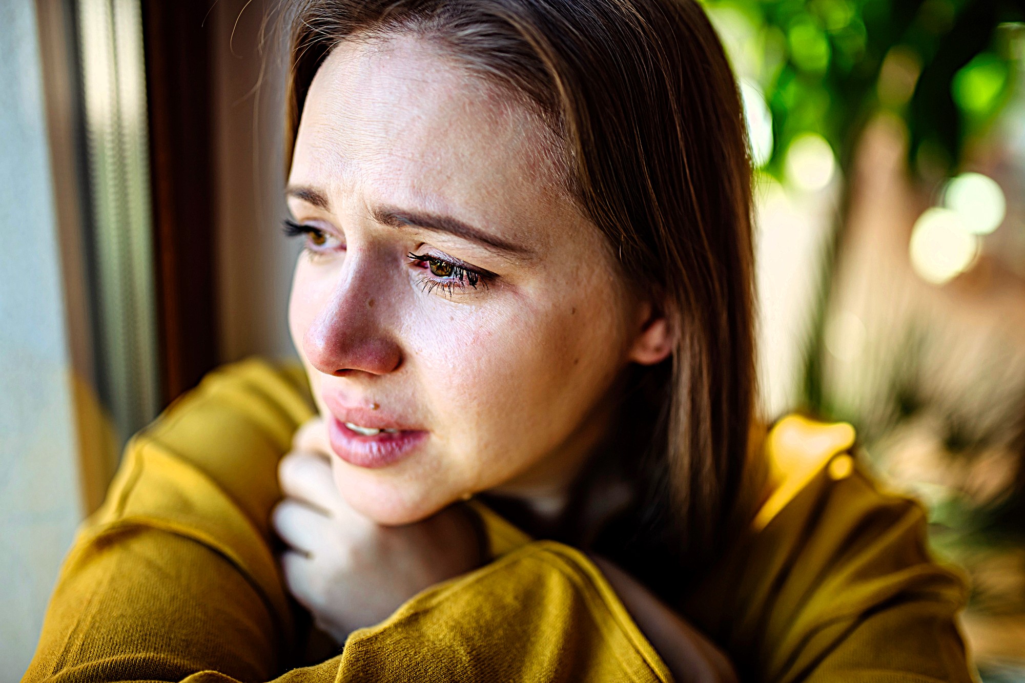 A woman with tearful eyes gazes out of a window, wearing a yellow sweater. She appears deep in thought, with sunlight illuminating her face and a blurred green background outside.