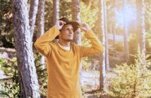 A person in a yellow sweater and black hat stands in a sunlit forest, adjusting their hat and gazing upwards. Tall trees surround them, and sunlight filters through the branches, creating a warm, serene atmosphere.