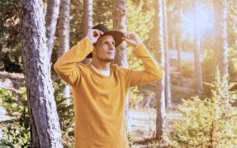 A person in a yellow sweater and black hat stands in a sunlit forest, adjusting their hat and gazing upwards. Tall trees surround them, and sunlight filters through the branches, creating a warm, serene atmosphere.