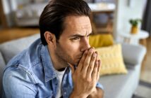 A man with dark hair and a denim shirt sits on a gray couch, appearing deep in thought with hands clasped near his mouth. A yellow pillow is in the background, adding a touch of color to the room.