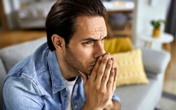 A man with dark hair and a denim shirt sits on a gray couch, appearing deep in thought with hands clasped near his mouth. A yellow pillow is in the background, adding a touch of color to the room.