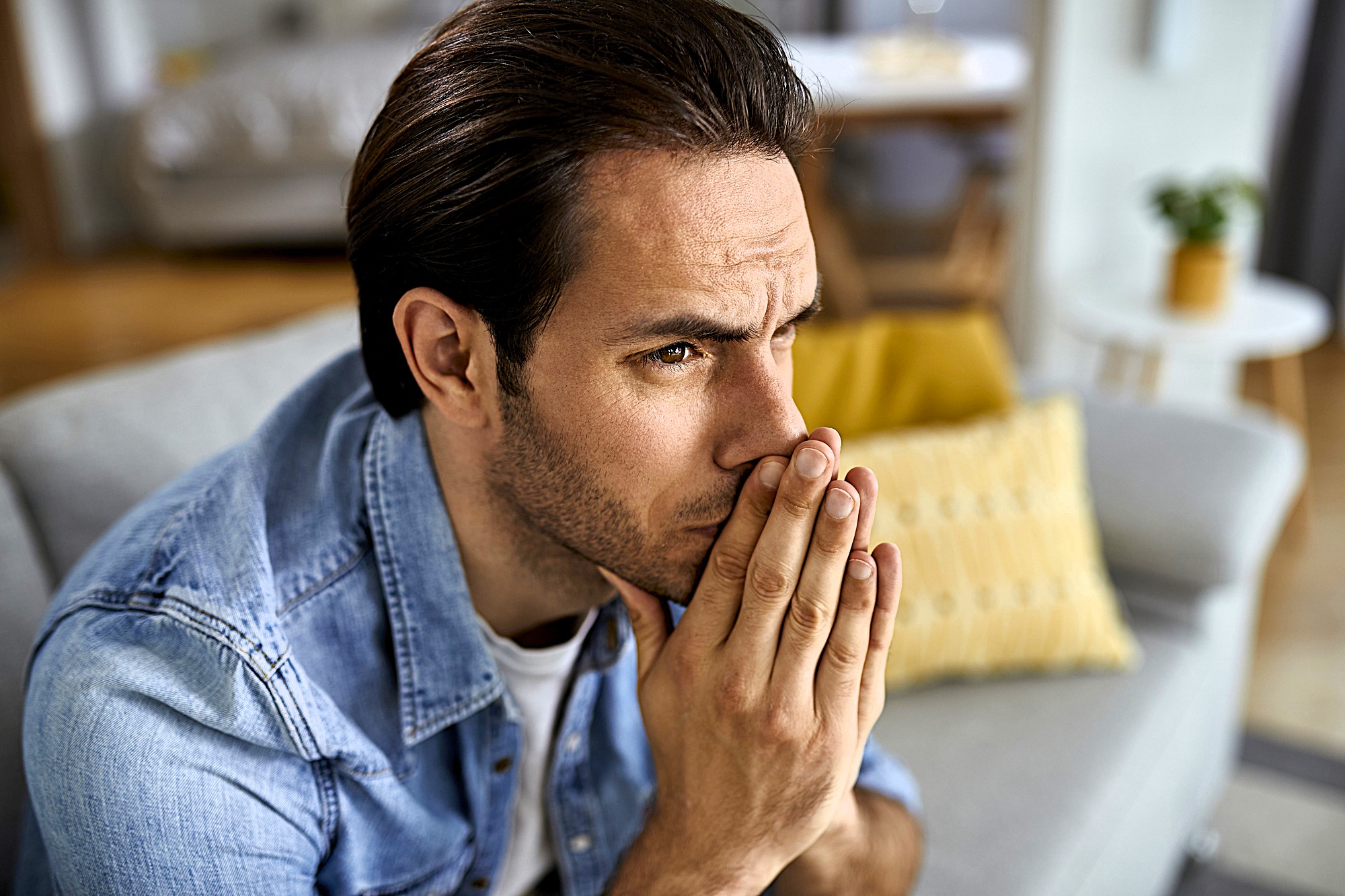 A man with dark hair and a denim shirt sits on a gray couch, appearing deep in thought with hands clasped near his mouth. A yellow pillow is in the background, adding a touch of color to the room.