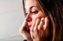 A woman with long brown hair rests her face on her hands, looking thoughtful and pensive. She is wearing a ring and gazing off to the side against a neutral background.