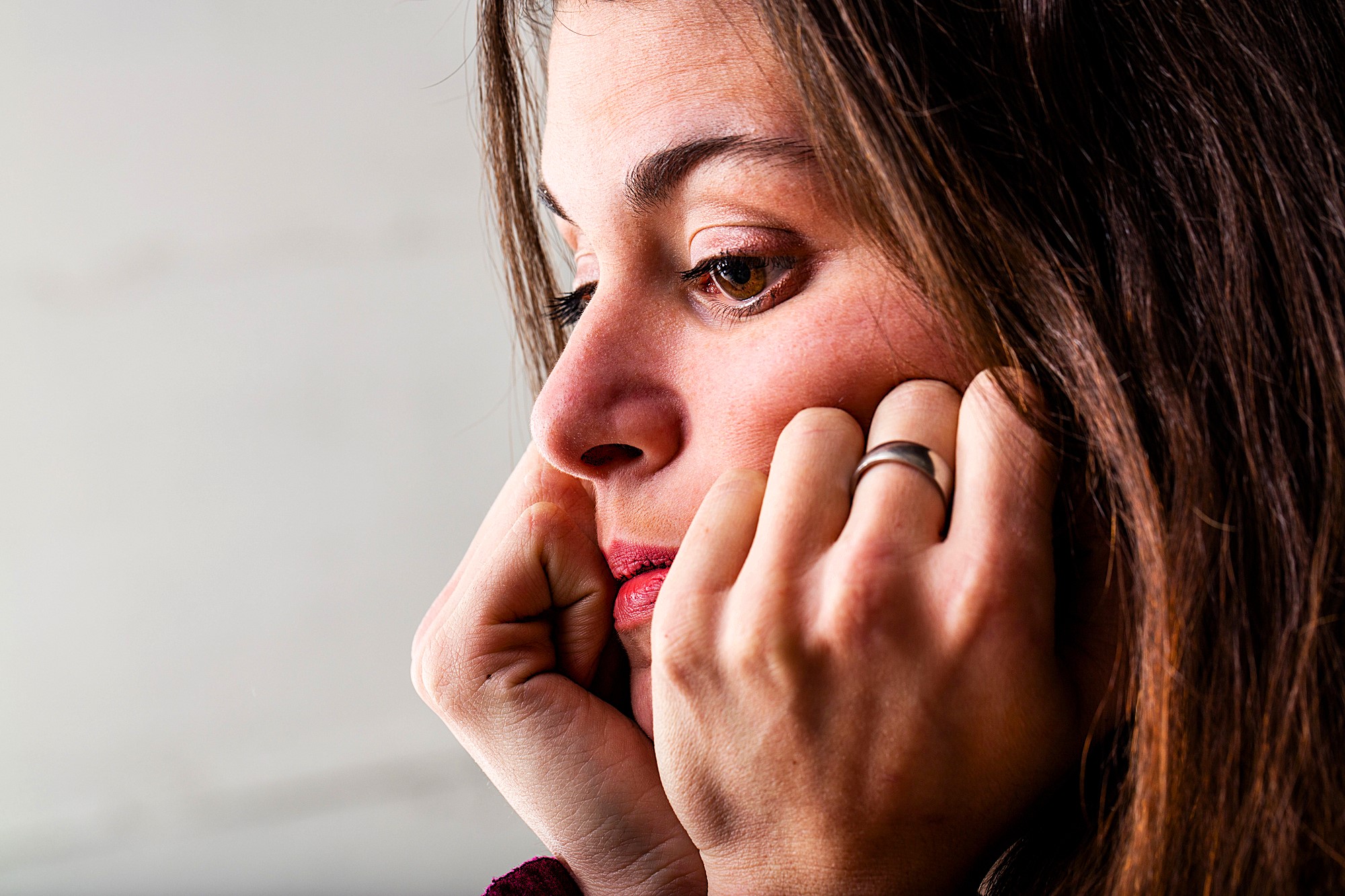 A woman with long brown hair rests her face on her hands, looking thoughtful and pensive. She is wearing a ring and gazing off to the side against a neutral background.