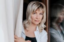 A woman with shoulder-length blonde hair and bangs stands by a window, looking directly at the camera. She is wearing a white shirt over a black top and gently holds a curtain aside, with her reflection visible in the glass.