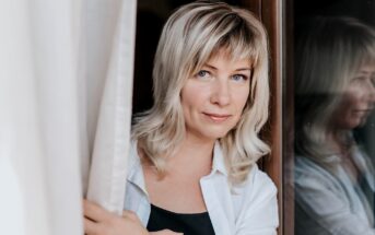 A woman with shoulder-length blonde hair and bangs stands by a window, looking directly at the camera. She is wearing a white shirt over a black top and gently holds a curtain aside, with her reflection visible in the glass.