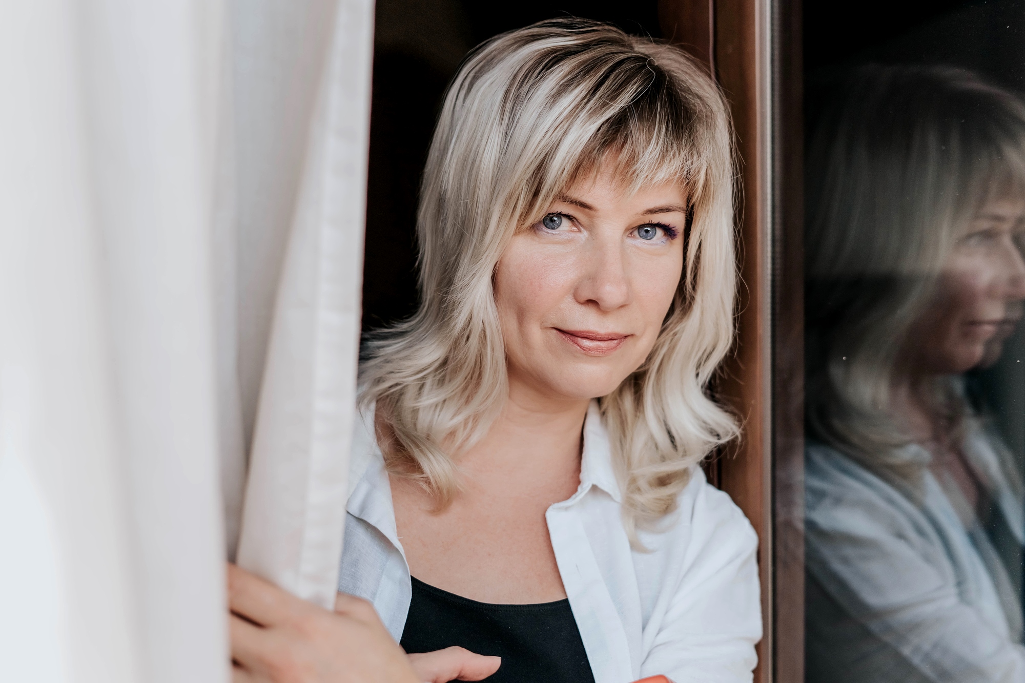 A woman with shoulder-length blonde hair and bangs stands by a window, looking directly at the camera. She is wearing a white shirt over a black top and gently holds a curtain aside, with her reflection visible in the glass.