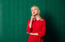 A woman in a red dress with a white collar stands thoughtfully against a green paneled background. She rests her chin on her hand and gazes upward, appearing contemplative.