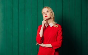 A woman in a red dress with a white collar stands thoughtfully against a green paneled background. She rests her chin on her hand and gazes upward, appearing contemplative.