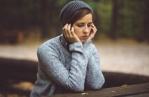 A person wearing a gray sweater and knitted hat sits pensively at a wooden table in a wooded area, resting their hands on their cheeks and gazing downward. The background is softly blurred with autumn leaves on the ground.