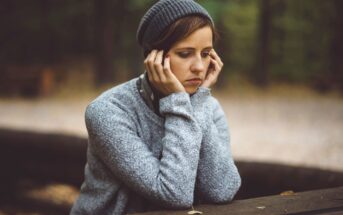 A person wearing a gray sweater and knitted hat sits pensively at a wooden table in a wooded area, resting their hands on their cheeks and gazing downward. The background is softly blurred with autumn leaves on the ground.