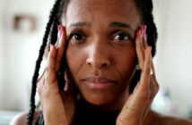 A person with long braids is holding their face with both hands, looking directly at the camera. The background is softly blurred, focusing attention on the person's expression.