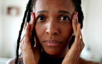 A person with long braids is holding their face with both hands, looking directly at the camera. The background is softly blurred, focusing attention on the person's expression.