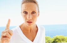 A person with short hair and a serious expression stands outdoors, raising their finger. They are wearing a white shirt, and the background features a clear blue sky and the ocean with some greenery.