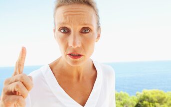 A person with short hair and a serious expression stands outdoors, raising their finger. They are wearing a white shirt, and the background features a clear blue sky and the ocean with some greenery.