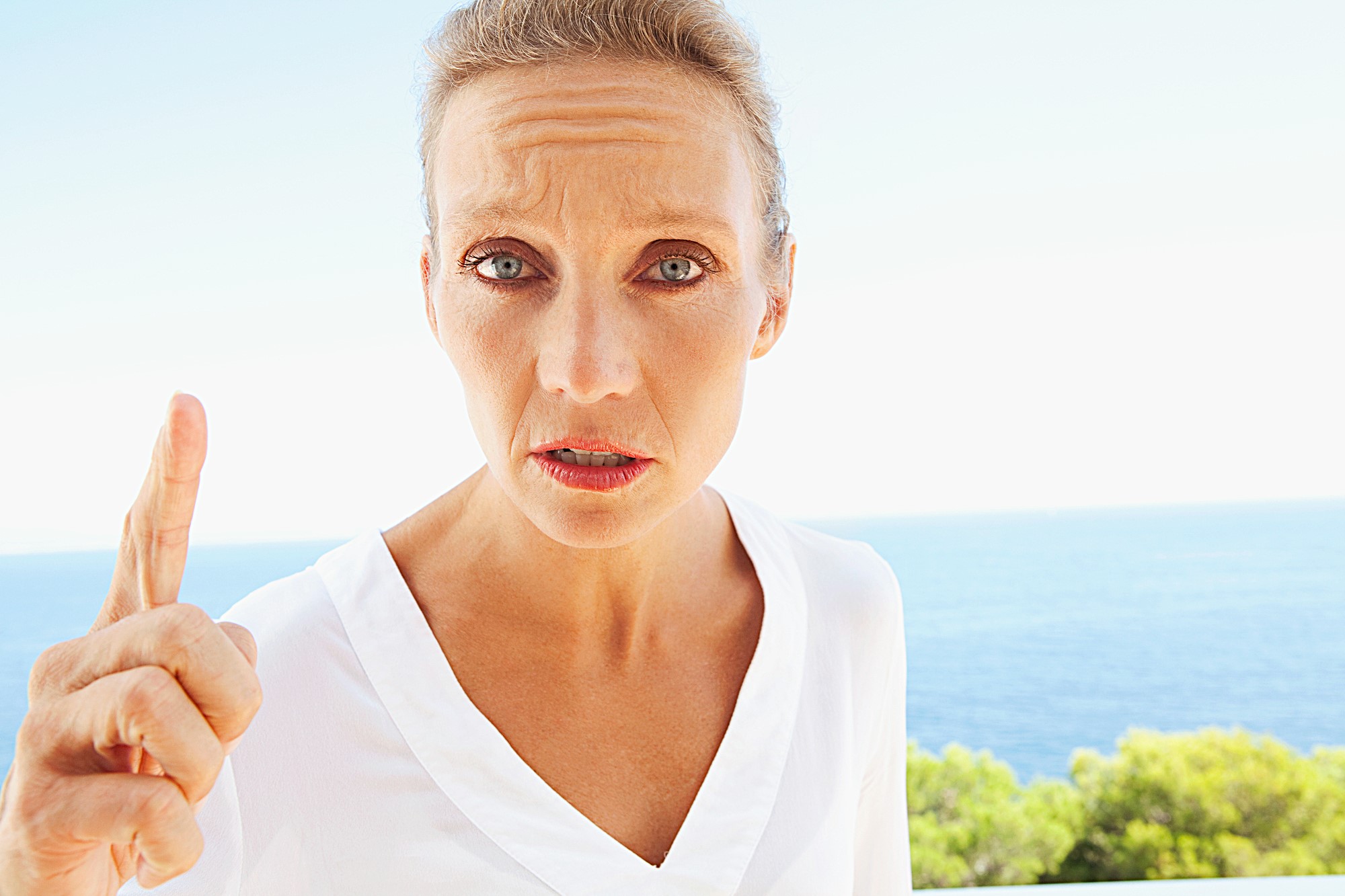 A person with short hair and a serious expression stands outdoors, raising their finger. They are wearing a white shirt, and the background features a clear blue sky and the ocean with some greenery.