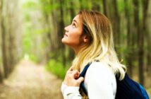 A woman with long blonde hair, wearing a white sweater and a backpack, looks up while standing on a forest path. Tall trees with green leaves surround her, creating a serene and natural setting.