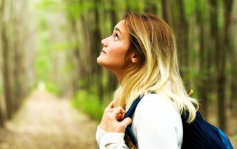 A woman with long blonde hair, wearing a white sweater and a backpack, looks up while standing on a forest path. Tall trees with green leaves surround her, creating a serene and natural setting.