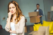 A woman with long brown hair looks thoughtful and concerned in the foreground. Behind her, a man carries moving boxes in a room with yellow cushions on the floor. The scene suggests a moving or packing scenario.