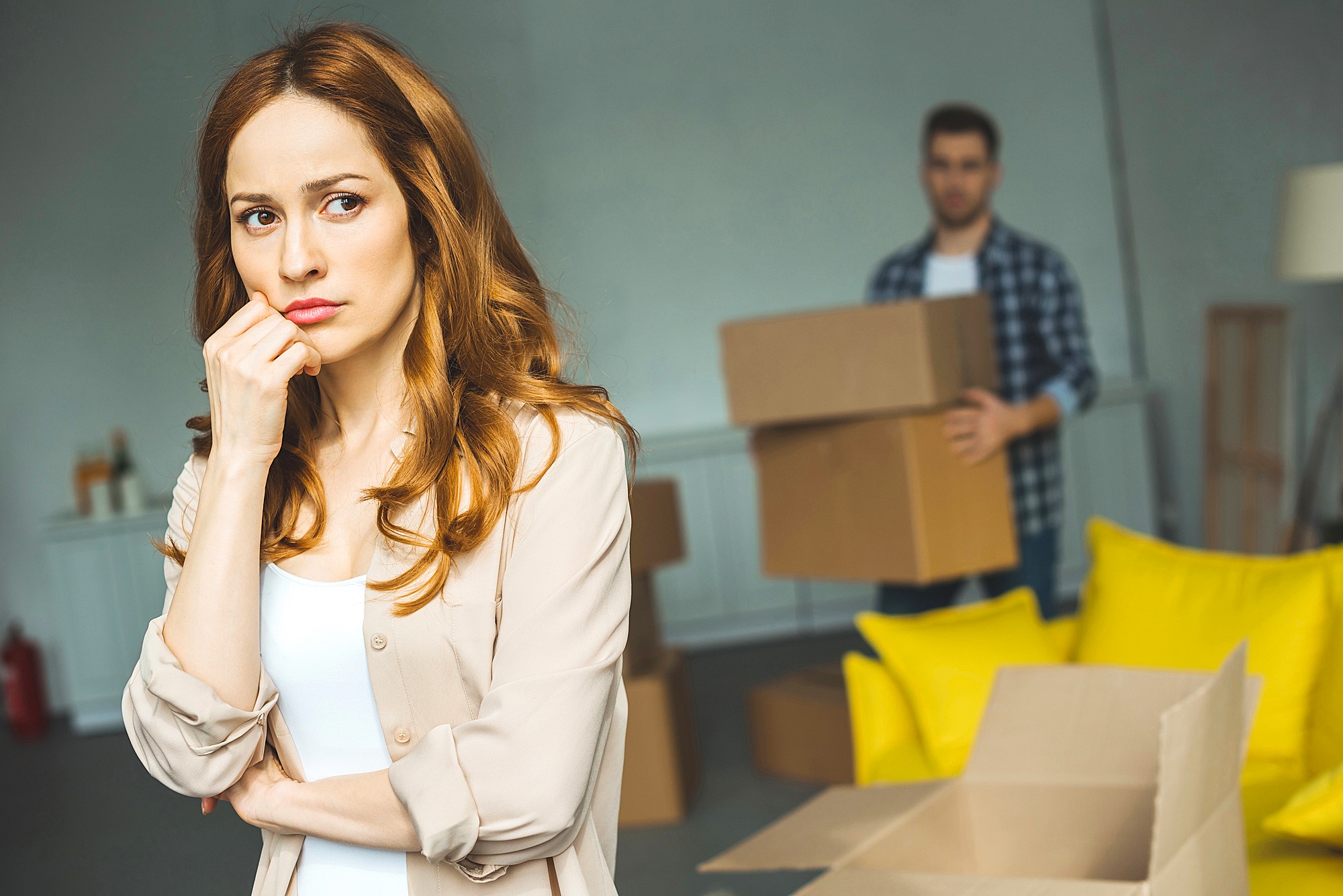 A woman with long brown hair looks thoughtful and concerned in the foreground. Behind her, a man carries moving boxes in a room with yellow cushions on the floor. The scene suggests a moving or packing scenario.