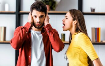 A man covers his ears with his hands, looking distressed, while a woman in a yellow top appears to be shouting at him. They are standing indoors in front of shelves lined with books and decor items.