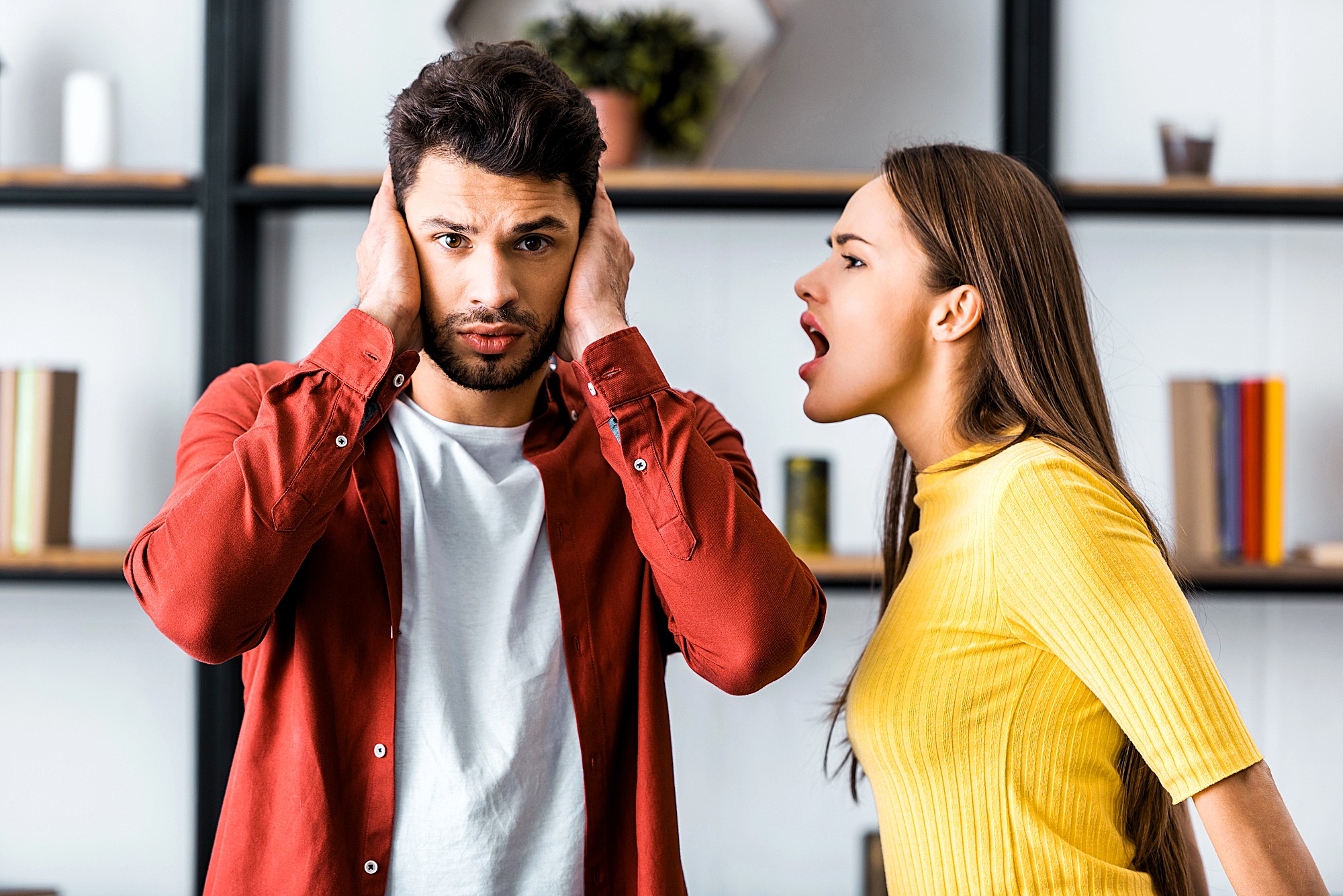 A man covers his ears with his hands, looking distressed, while a woman in a yellow top appears to be shouting at him. They are standing indoors in front of shelves lined with books and decor items.