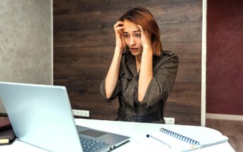 A worried woman sits at a table with her hands on her forehead, looking at a laptop screen. A notebook and pen are on the table beside her. The background features a modern wood-paneled wall.