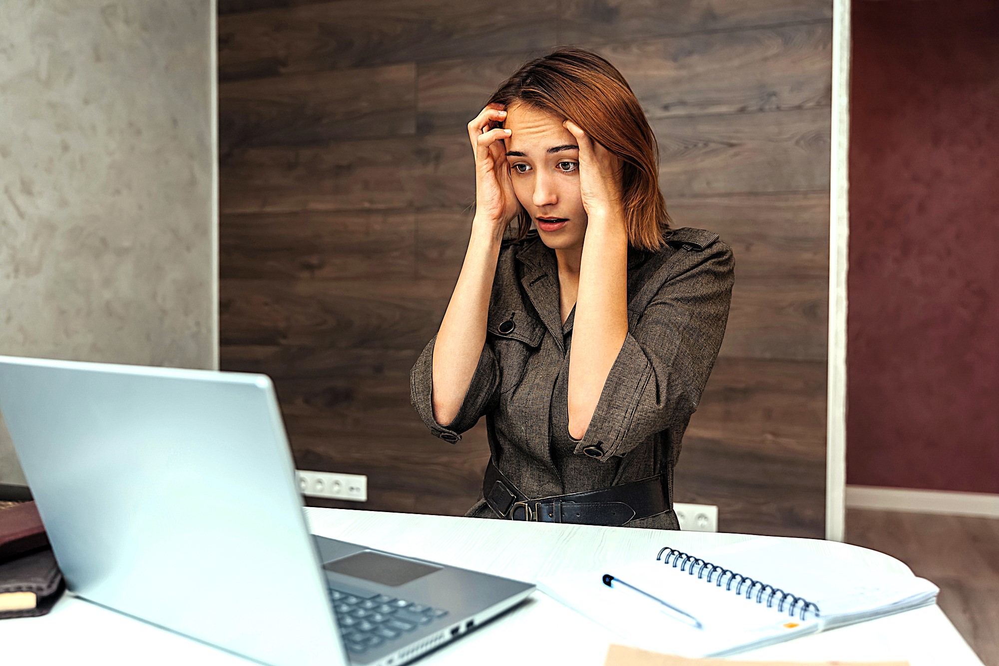 A worried woman sits at a table with her hands on her forehead, looking at a laptop screen. A notebook and pen are on the table beside her. The background features a modern wood-paneled wall.