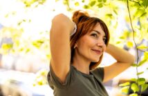 A woman with short red hair stands outdoors, smiling while adjusting her hair with both hands. She is wearing a gray shirt, surrounded by green leaves and sunlight filtering through the trees. The background is softly blurred.