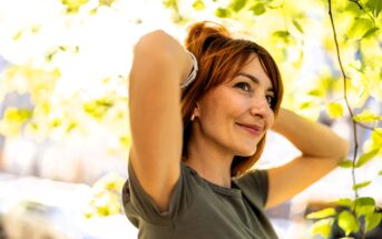 A woman with short red hair stands outdoors, smiling while adjusting her hair with both hands. She is wearing a gray shirt, surrounded by green leaves and sunlight filtering through the trees. The background is softly blurred.