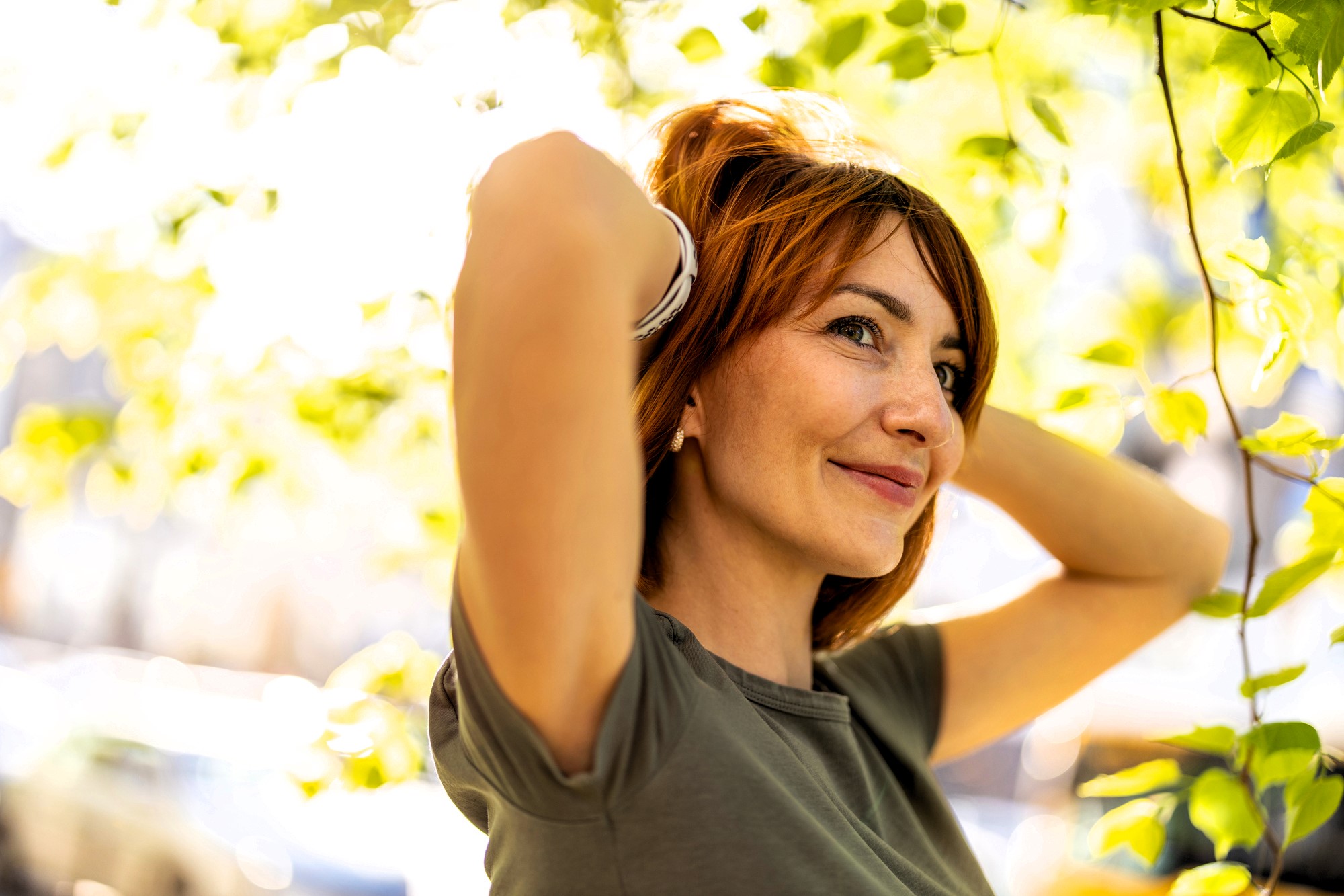 A woman with short red hair stands outdoors, smiling while adjusting her hair with both hands. She is wearing a gray shirt, surrounded by green leaves and sunlight filtering through the trees. The background is softly blurred.