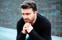 A man with a thoughtful expression sits outdoors against a textured dark wall, resting his chin on his hands. He wears a black sweater and has short brown hair and a beard.