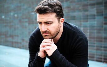 A man with a thoughtful expression sits outdoors against a textured dark wall, resting his chin on his hands. He wears a black sweater and has short brown hair and a beard.