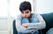 A person with short dark hair sits on a couch, wearing a gray sweater and blue jeans, looking directly at the camera with a serious expression. They are hugging their knees, with natural light coming from a window behind them.