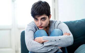 A person with short dark hair sits on a couch, wearing a gray sweater and blue jeans, looking directly at the camera with a serious expression. They are hugging their knees, with natural light coming from a window behind them.