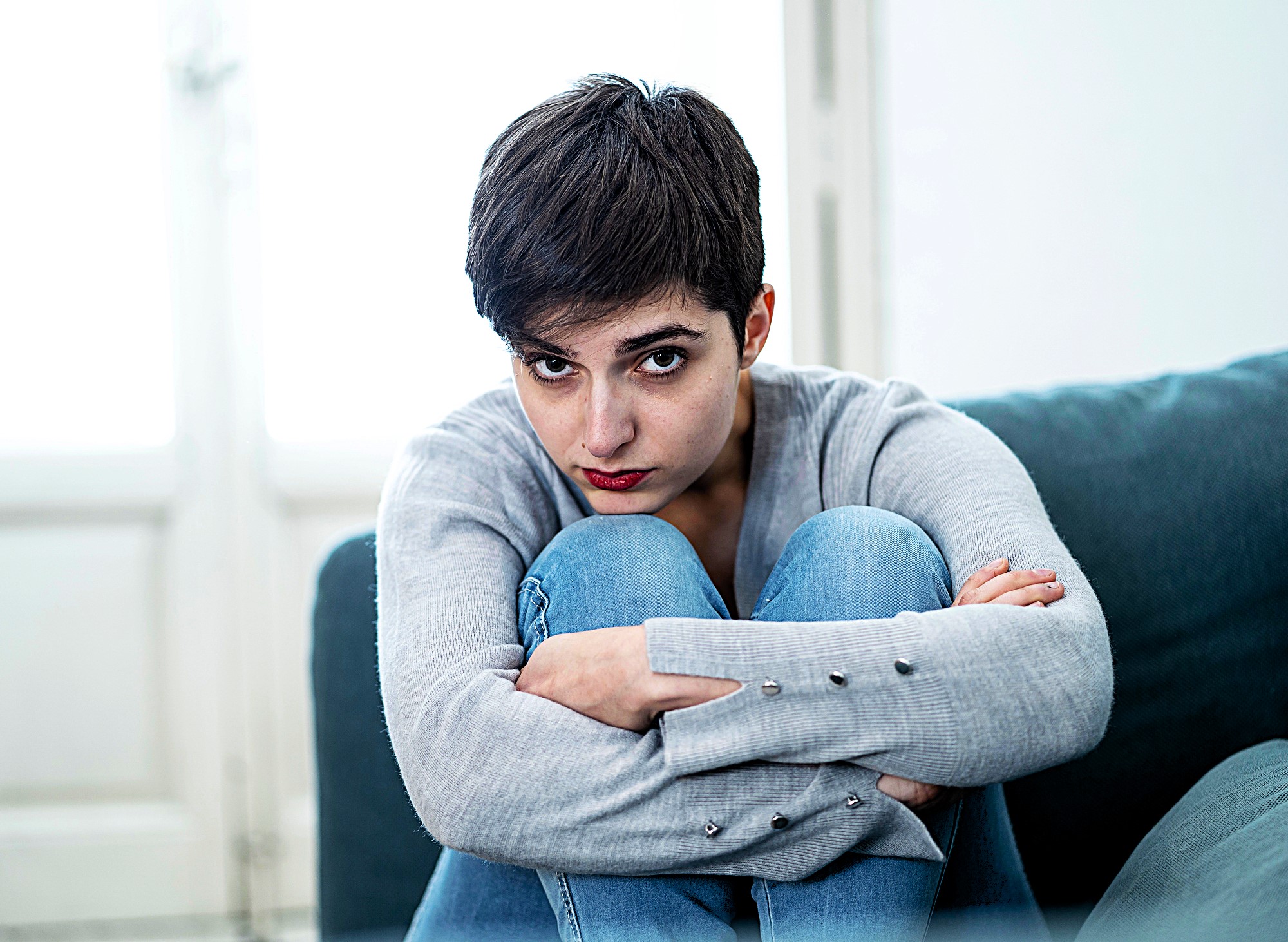 A person with short dark hair sits on a couch, wearing a gray sweater and blue jeans, looking directly at the camera with a serious expression. They are hugging their knees, with natural light coming from a window behind them.